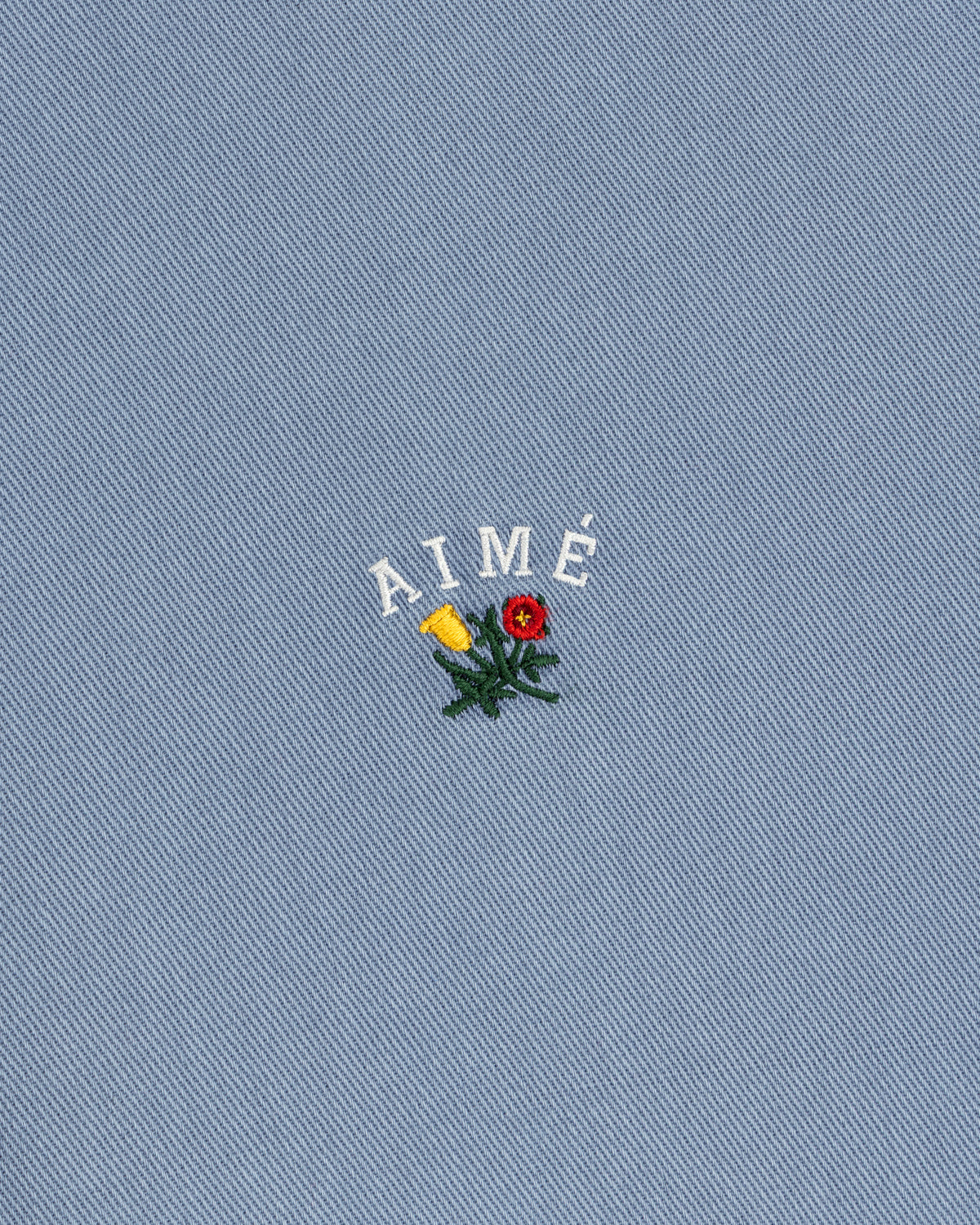 Garment Dyed Leon Overshirt: blue denim with AIMÉ text and embroidered red/yellow flowers with green leaves.