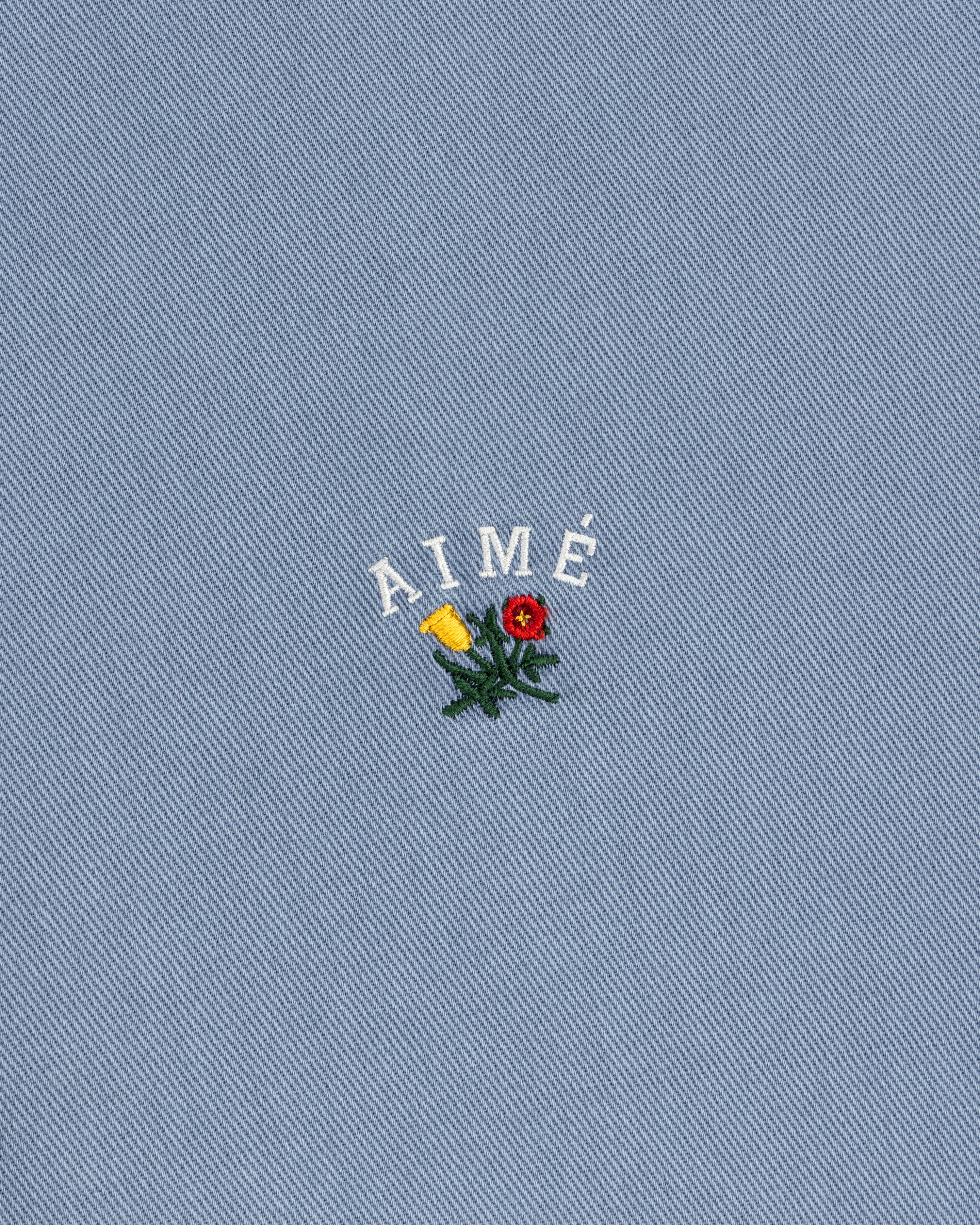 Garment Dyed Leon Overshirt: blue denim with AIMÉ text and embroidered red/yellow flowers with green leaves.
