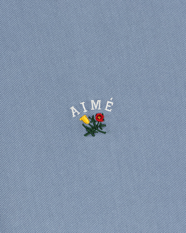 Garment Dyed Leon Overshirt: blue denim with AIMÉ text and embroidered red/yellow flowers with green leaves.
