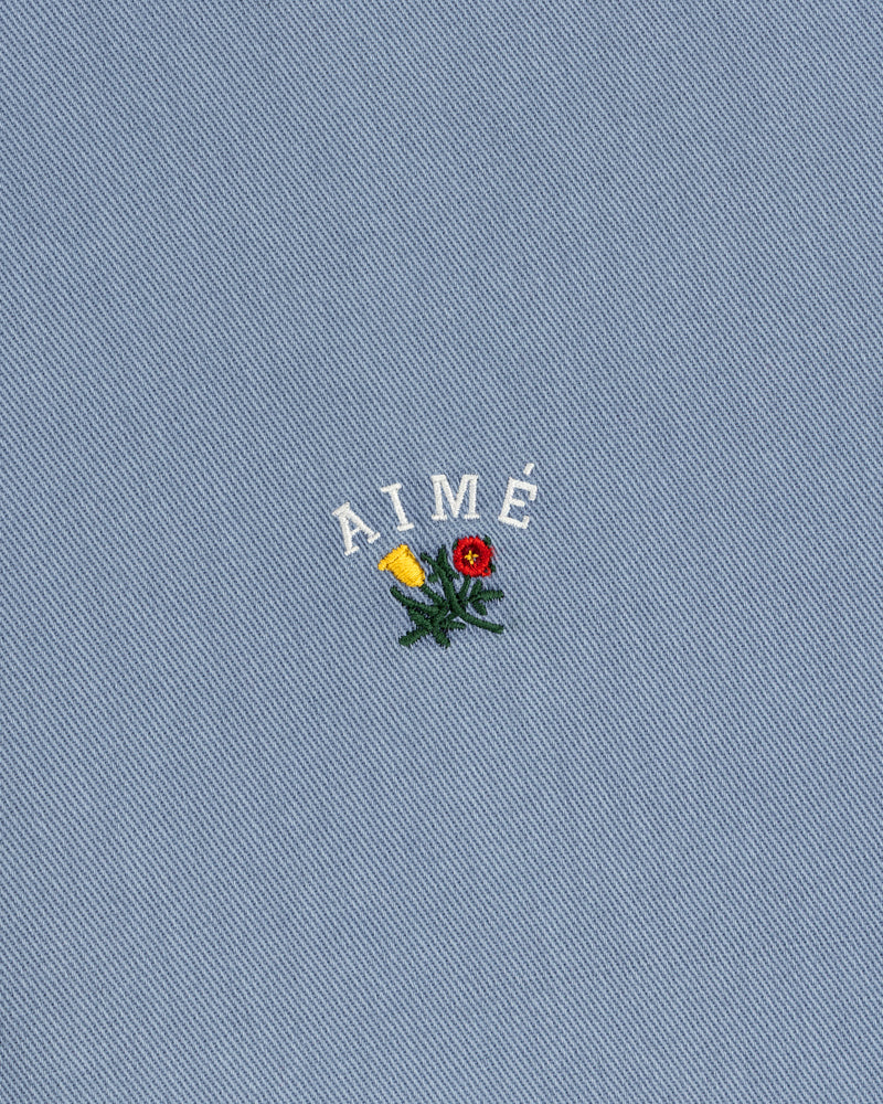 Garment Dyed Leon Overshirt: blue denim with AIMÉ text and embroidered red/yellow flowers with green leaves.