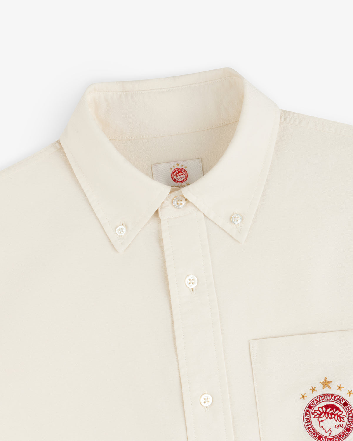 Cream-colored ALD / Olympiacos F.C. Logo Oxford Shirt with a red athletic logo on the chest pocket and inner collar tag.