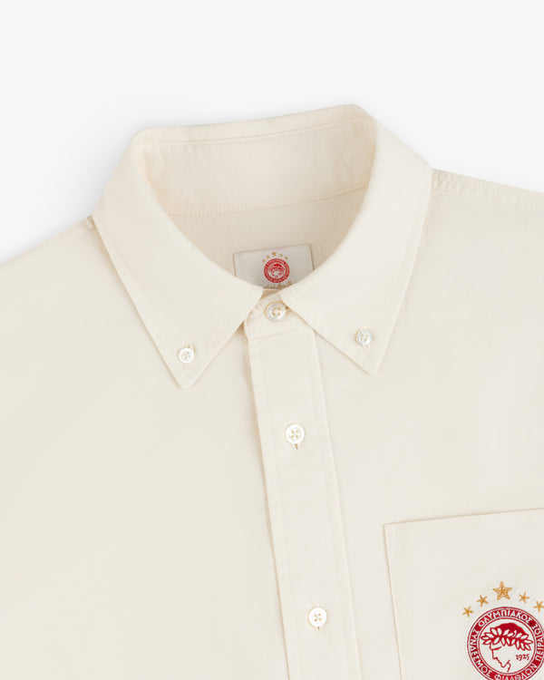 Cream-colored ALD / Olympiacos F.C. Logo Oxford Shirt with a red athletic logo on the chest pocket and inner collar tag.