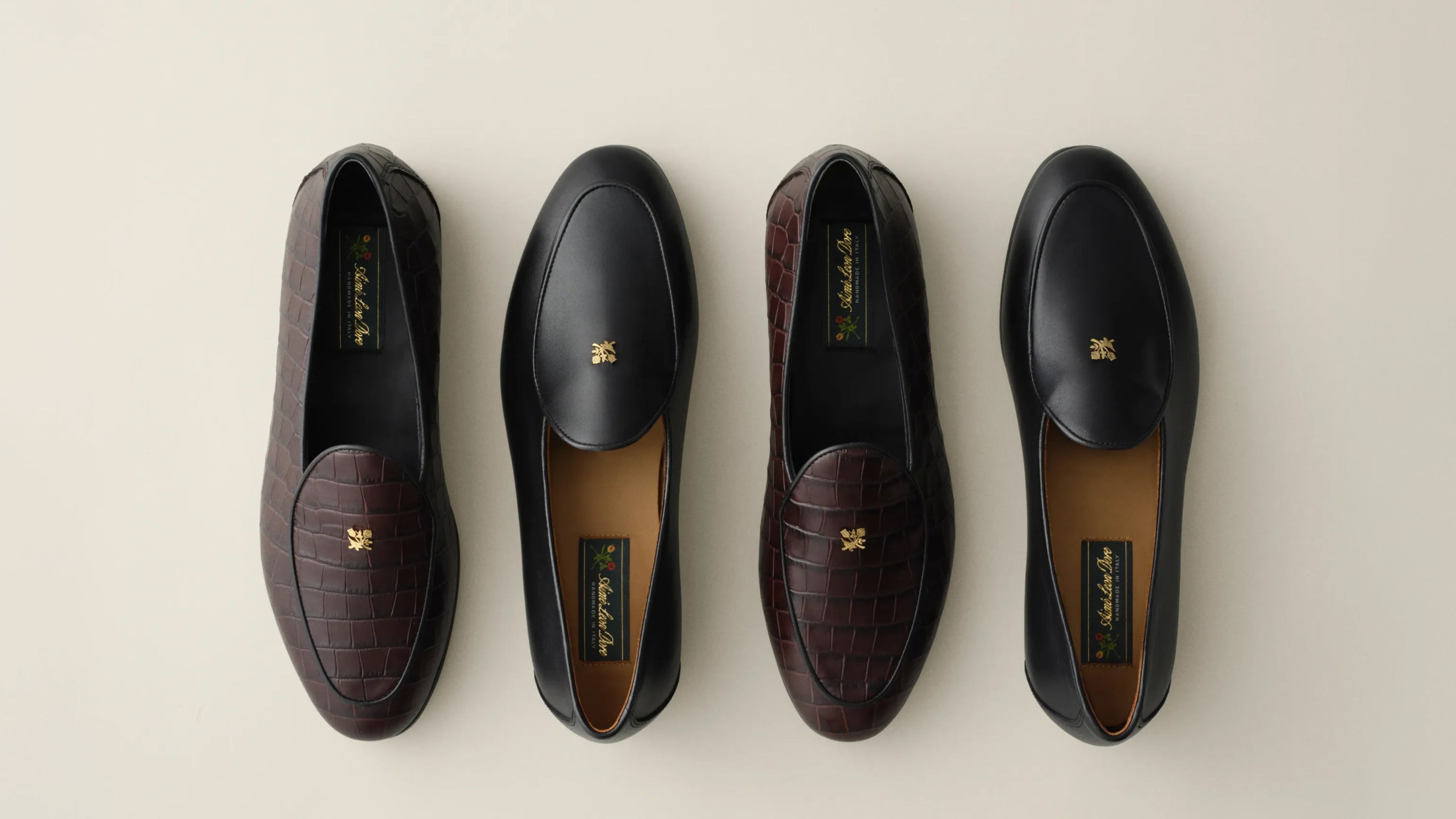 Four loafers arranged in a row on a light beige background, alternating between dark brown crocodile-embossed leather and black smooth leather, each with a small gold logo on the toe.