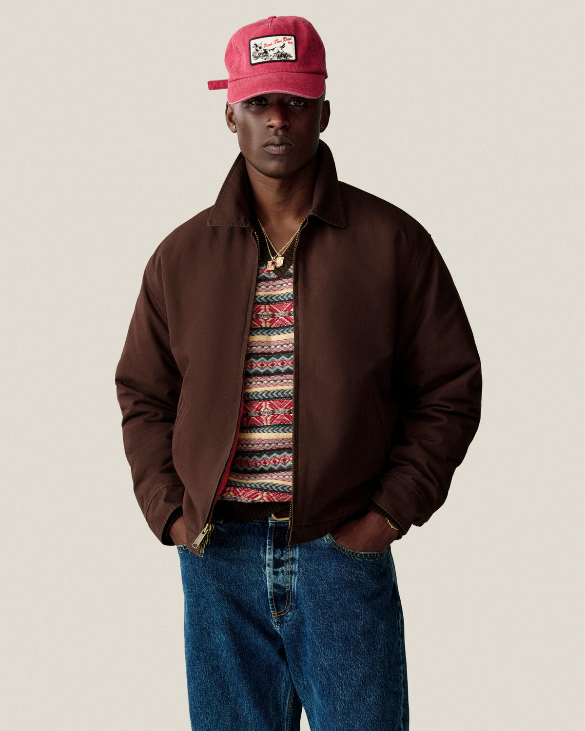 Man in brown jacket, Fair Isle V-Neck Sweater, jeans, and red cap stands hands-in-pockets against plain background.