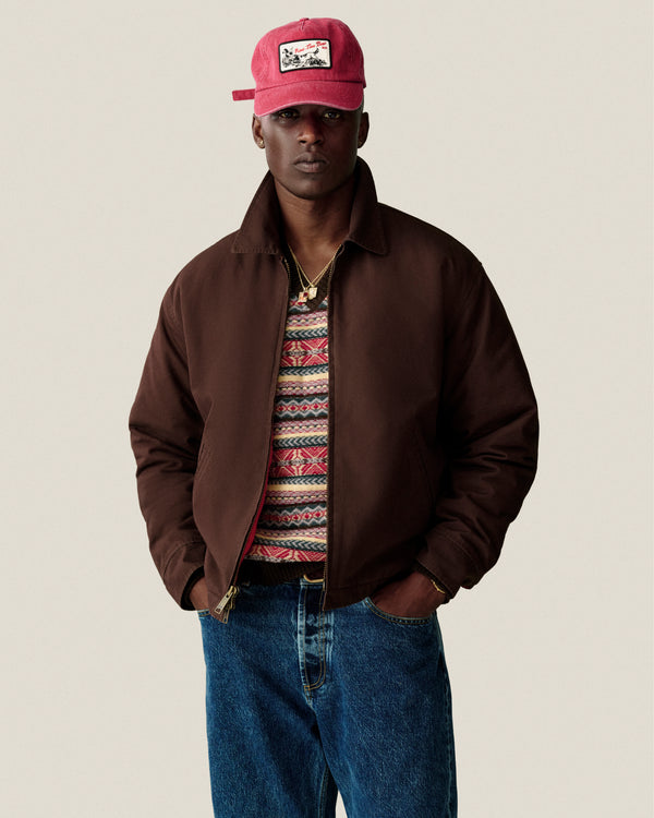 Man in brown jacket, Fair Isle V-Neck Sweater, jeans, and red cap stands hands-in-pockets against plain background.