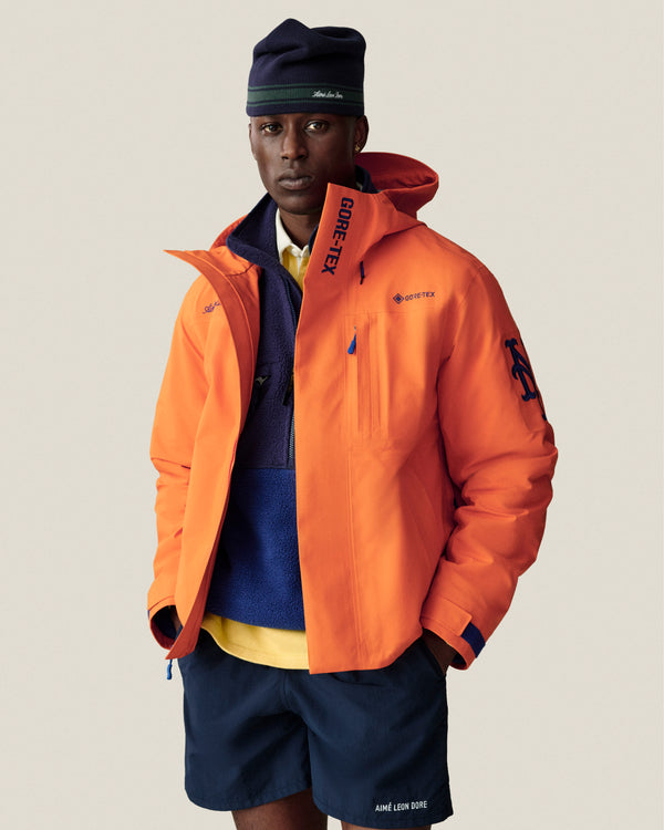 Man wearing an orange GORE-TEX jacket, blue fleece, navy shorts, and a Stripe Knit Beanie against a plain background.