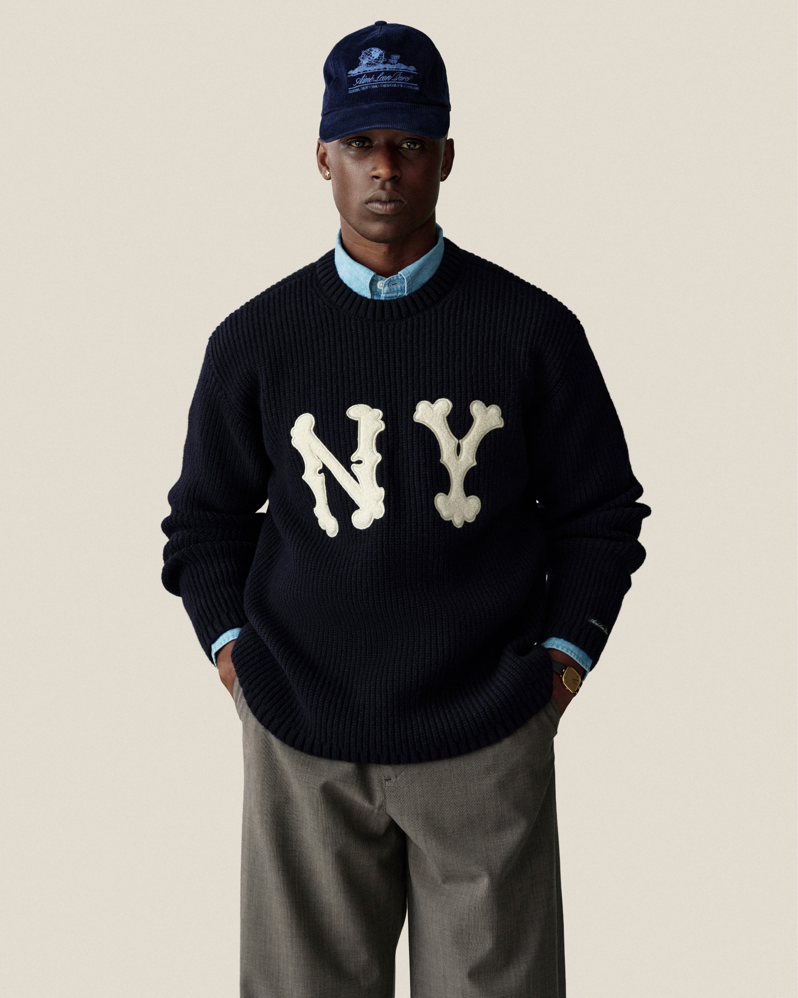 Man in navy cap, Bayard Chambray Shirt, black NY sweater, and gray pants against a beige background.