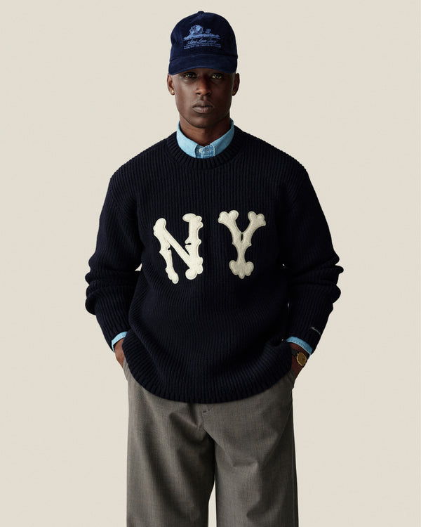 Man in navy cap, Bayard Chambray Shirt, black NY sweater, and gray pants against a beige background.