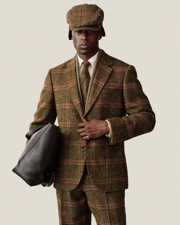 A man in a green and brown plaid tweed suit, matching cap, and Plaid Tweed Tie, holding a large leather bag.