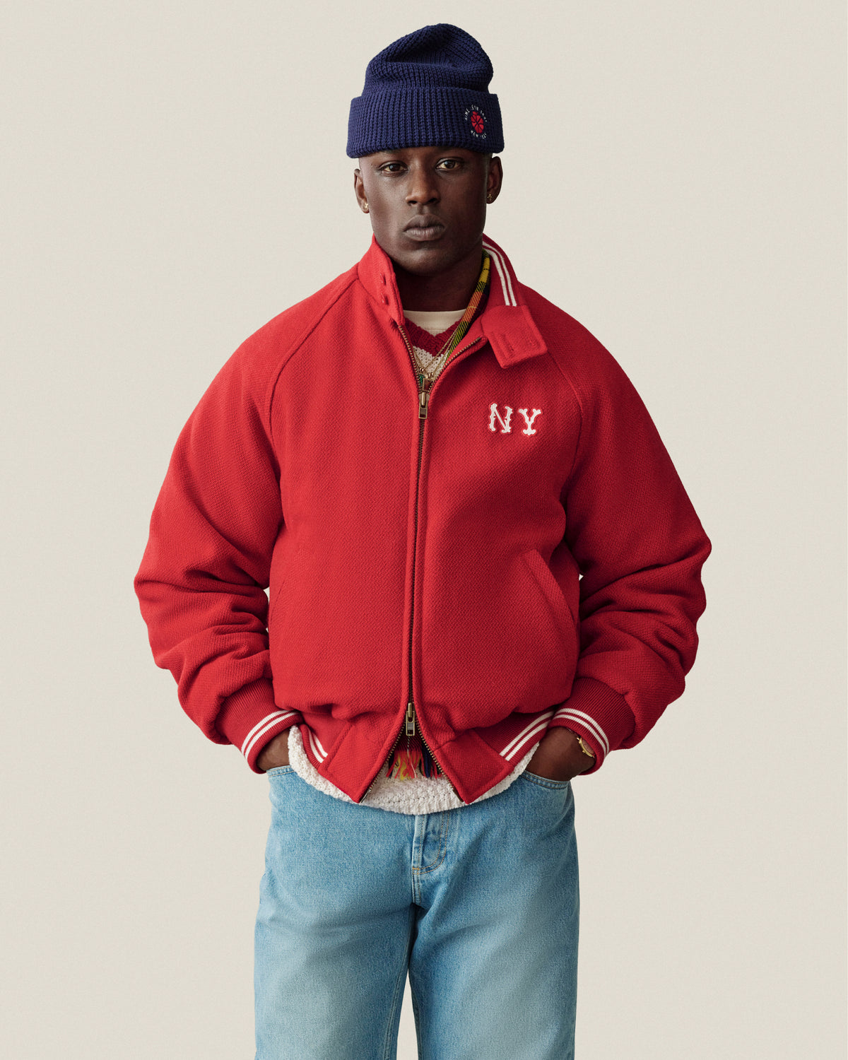 Someone in an NY Highlanders Wool Varsity Jacket, blue jeans, and a navy beanie stands by a beige background.
