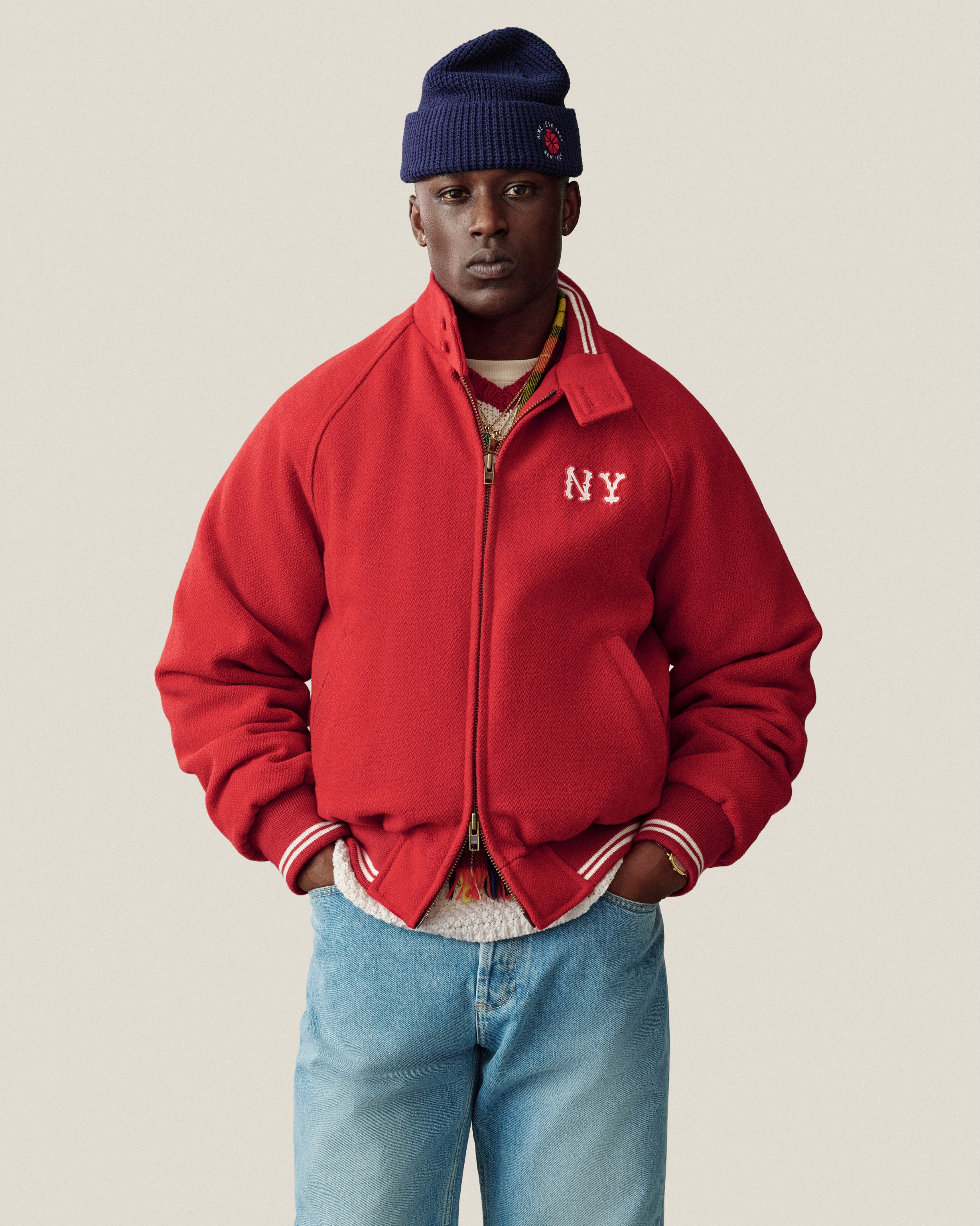 Someone in an NY Highlanders Wool Varsity Jacket, blue jeans, and a navy beanie stands by a beige background.