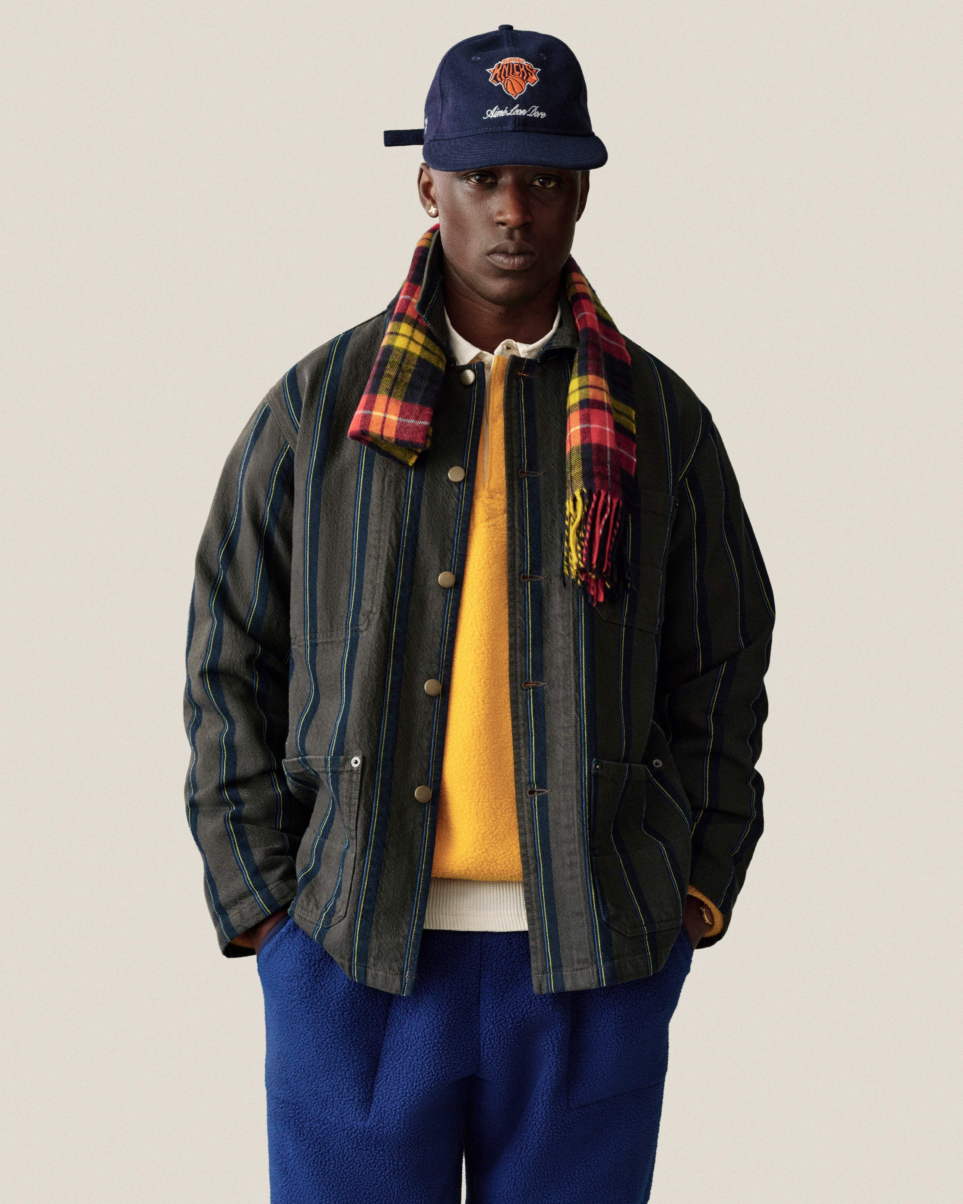 Man in Striped Denim Chore Jacket, yellow shirt, blue pants, plaid scarf & navy cap on plain background.