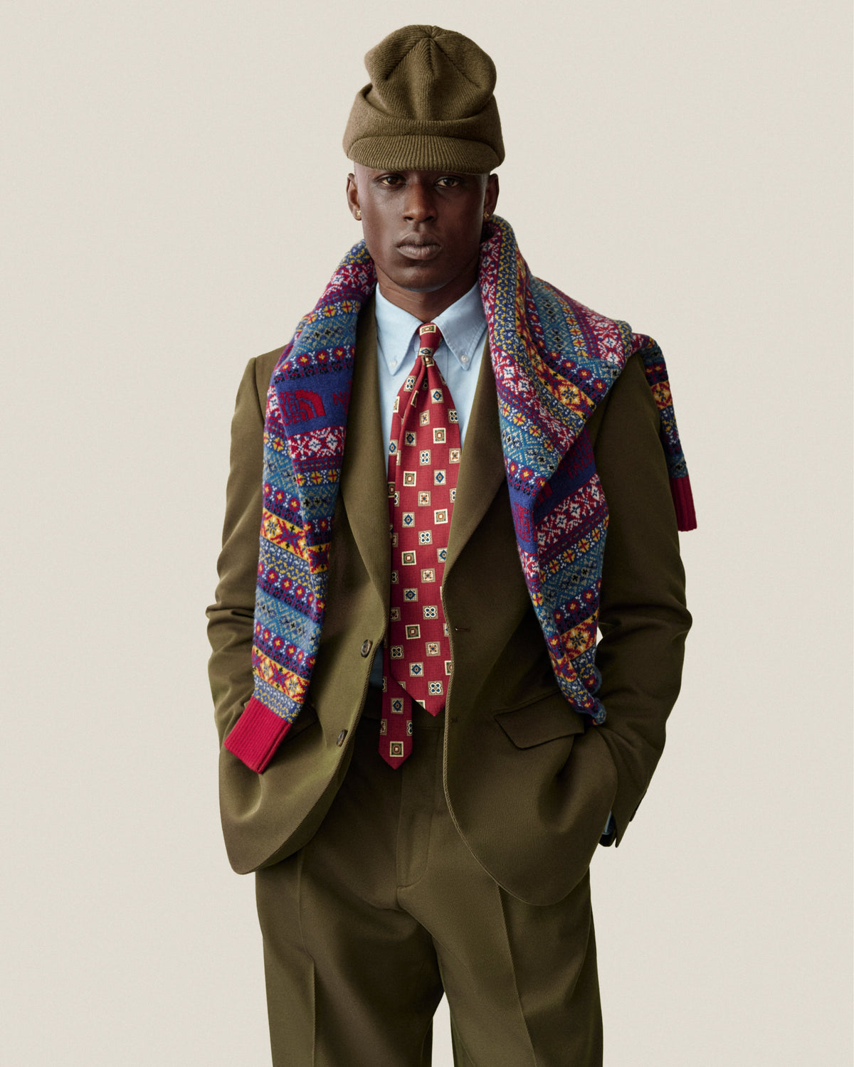 A man wearing an olive Single-Breasted Wool Blend Suit Jacket, blue shirt, patterned red tie, knit beanie, and scarf.