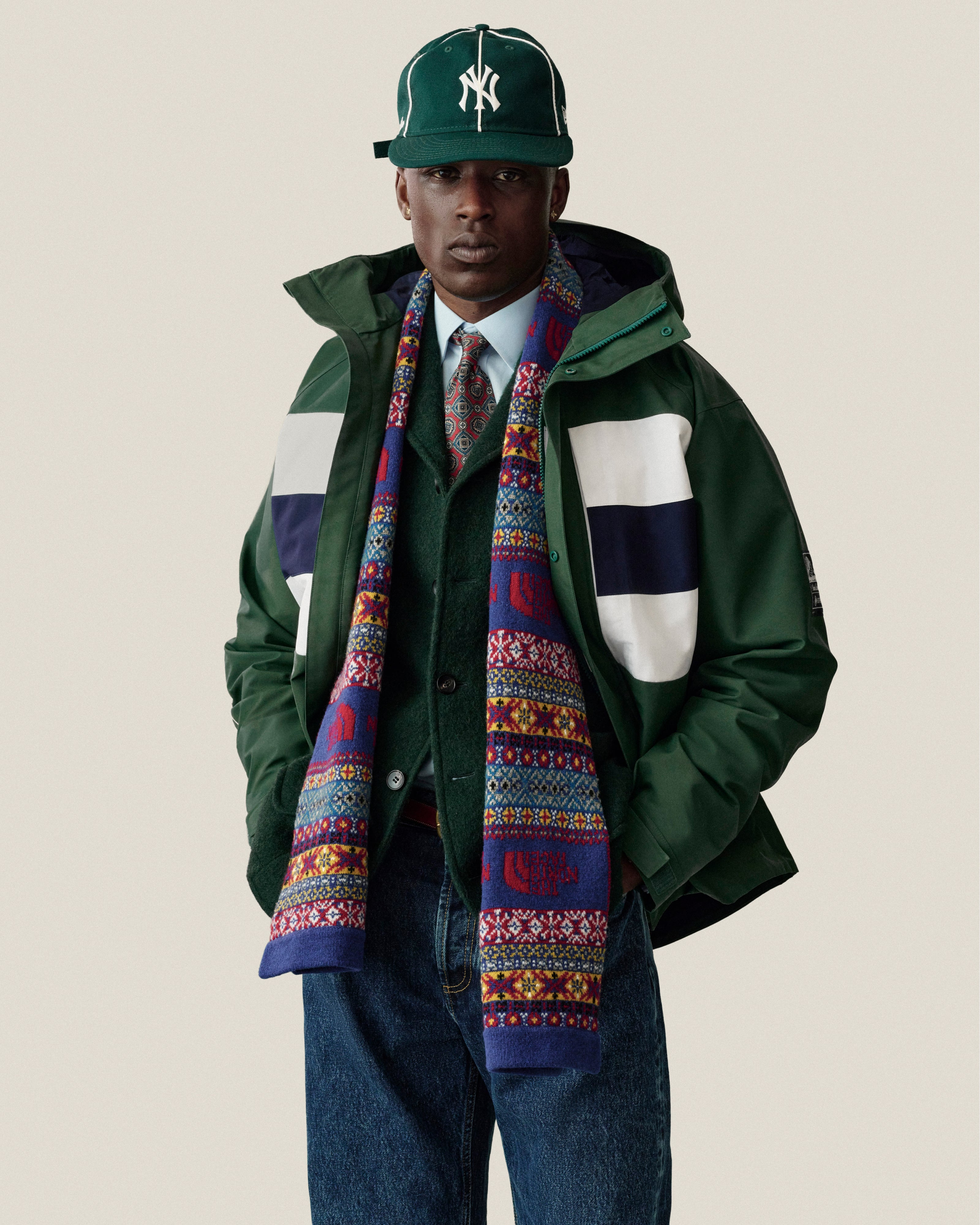 A man in a GORE-TEX Striped Sailing Jacket, NY cap, scarf, shirt, tie & jeans stands by a plain background.