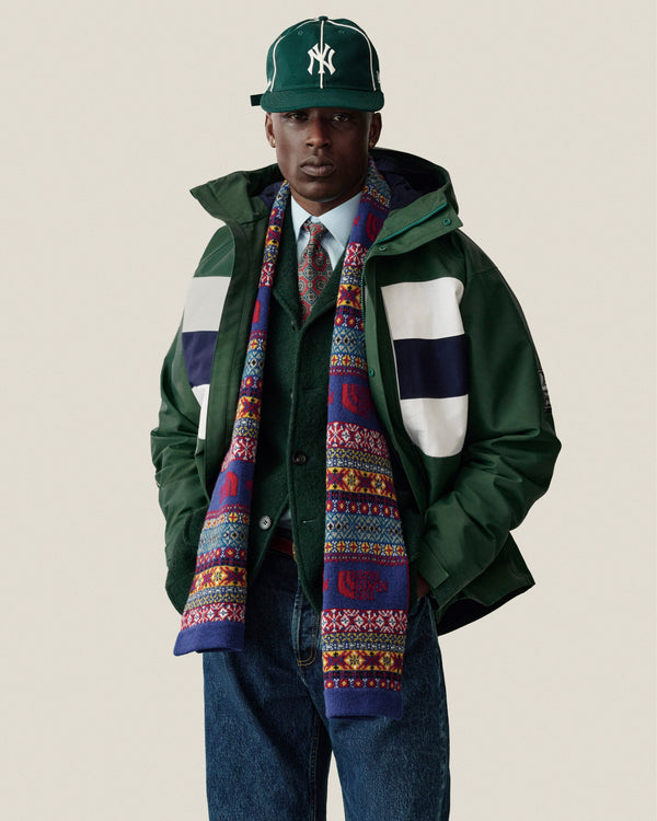 A man in a GORE-TEX Striped Sailing Jacket, NY cap, scarf, shirt, tie & jeans stands by a plain background.