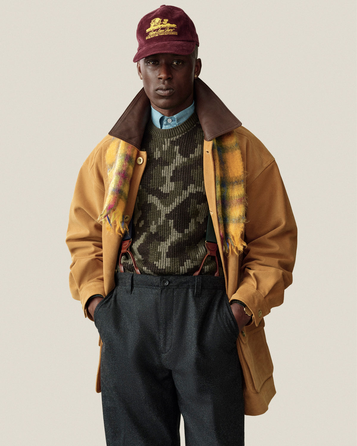 A person wearing a Wool Jacquard Camo Sweater under a tan jacket, a plaid scarf, and a maroon cap.