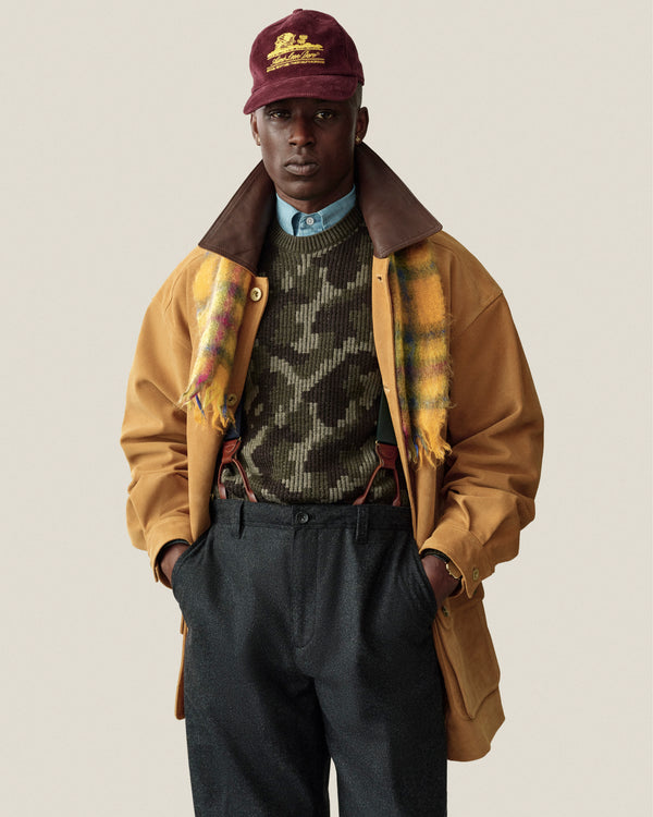 A person wearing a Wool Jacquard Camo Sweater under a tan jacket, a plaid scarf, and a maroon cap.