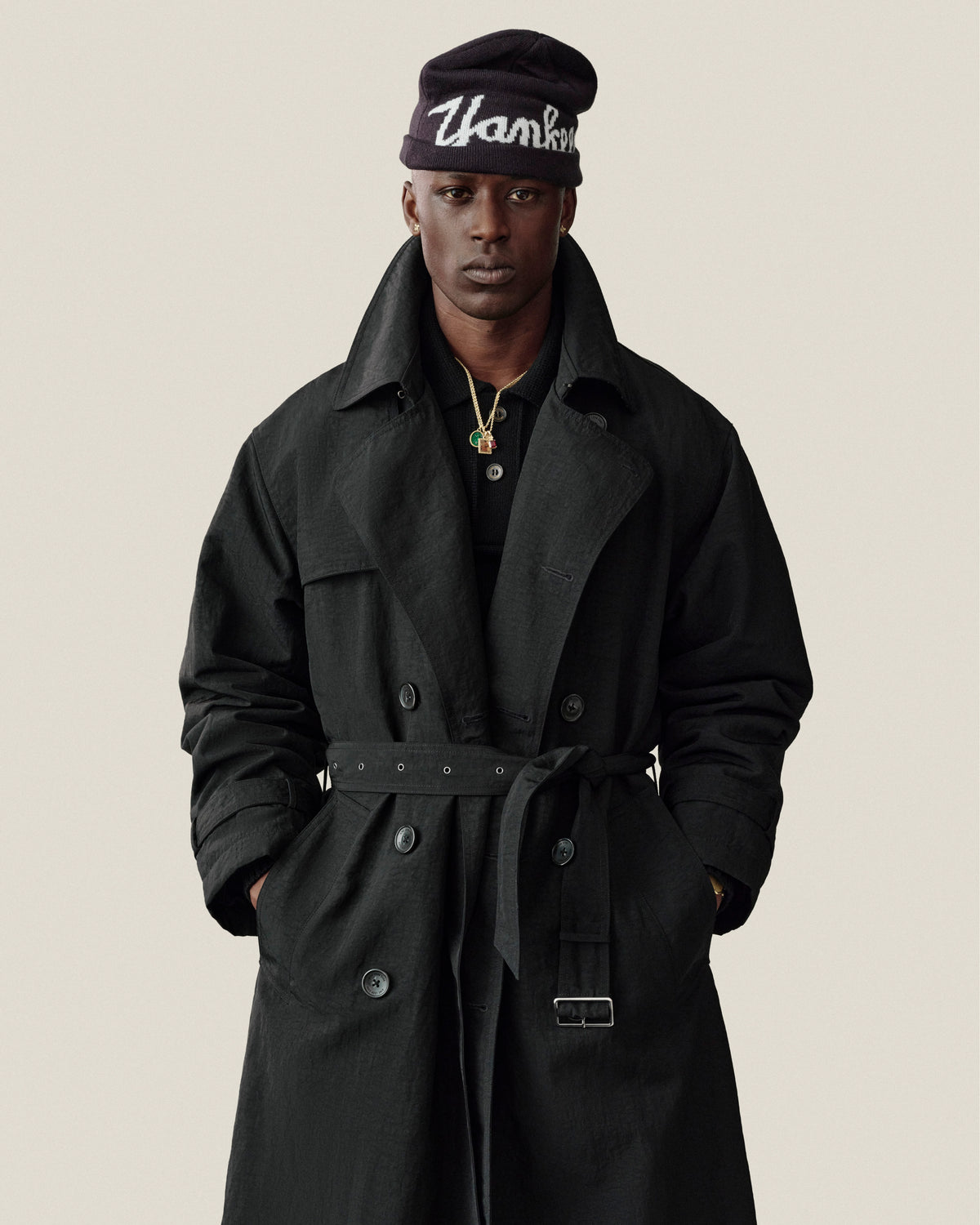 A person in a Double Breasted Nylon Trench Coat and “Thanks” beanie stands against a plain background.