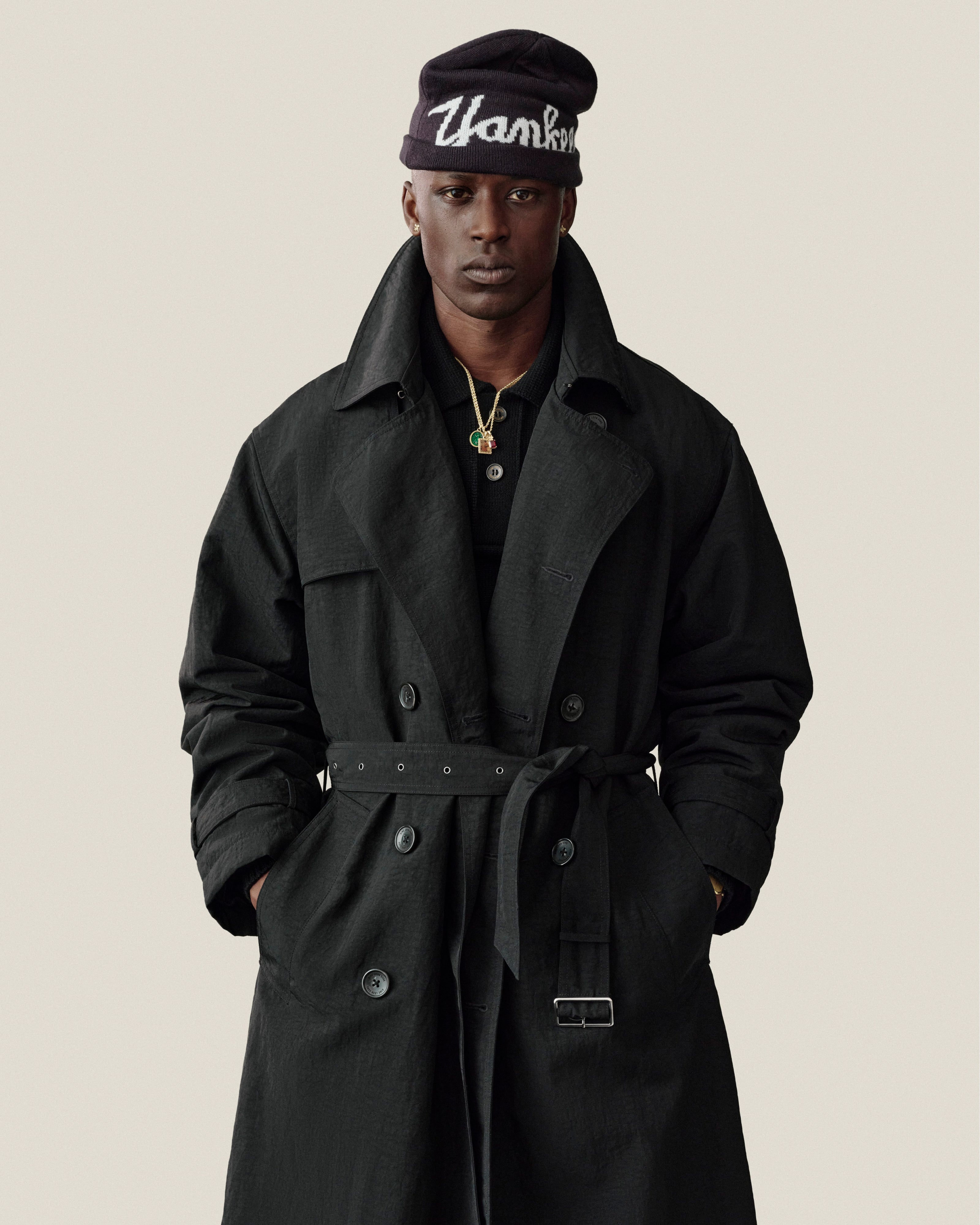 A person in a Double Breasted Nylon Trench Coat and “Thanks” beanie stands against a plain background.