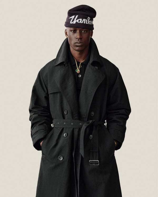 A person in a Double Breasted Nylon Trench Coat and “Thanks” beanie stands against a plain background.