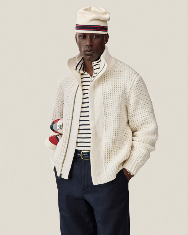 Man in Stripe Knit Beanie, cream jacket, striped shirt, holding rugby balls against a plain background.