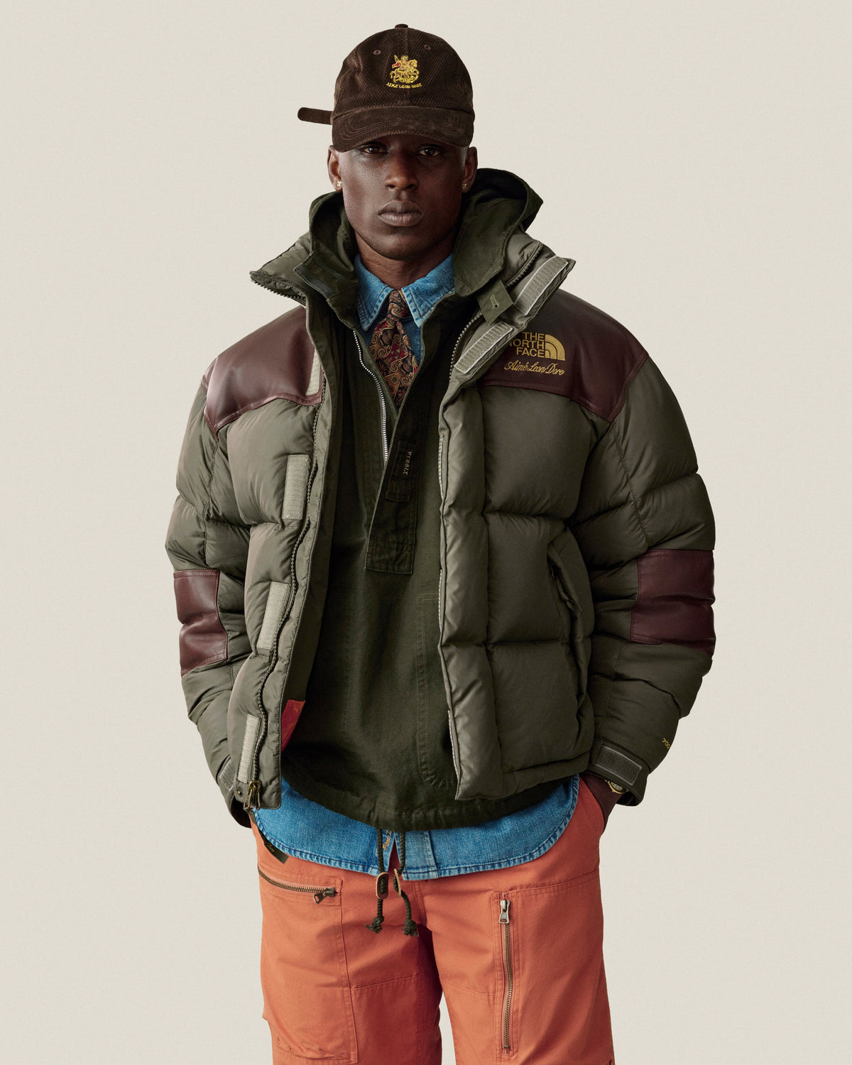 A man in a puffer jacket, Saint George Corduroy Hat, patterned tie, and orange pants stands against a plain background.