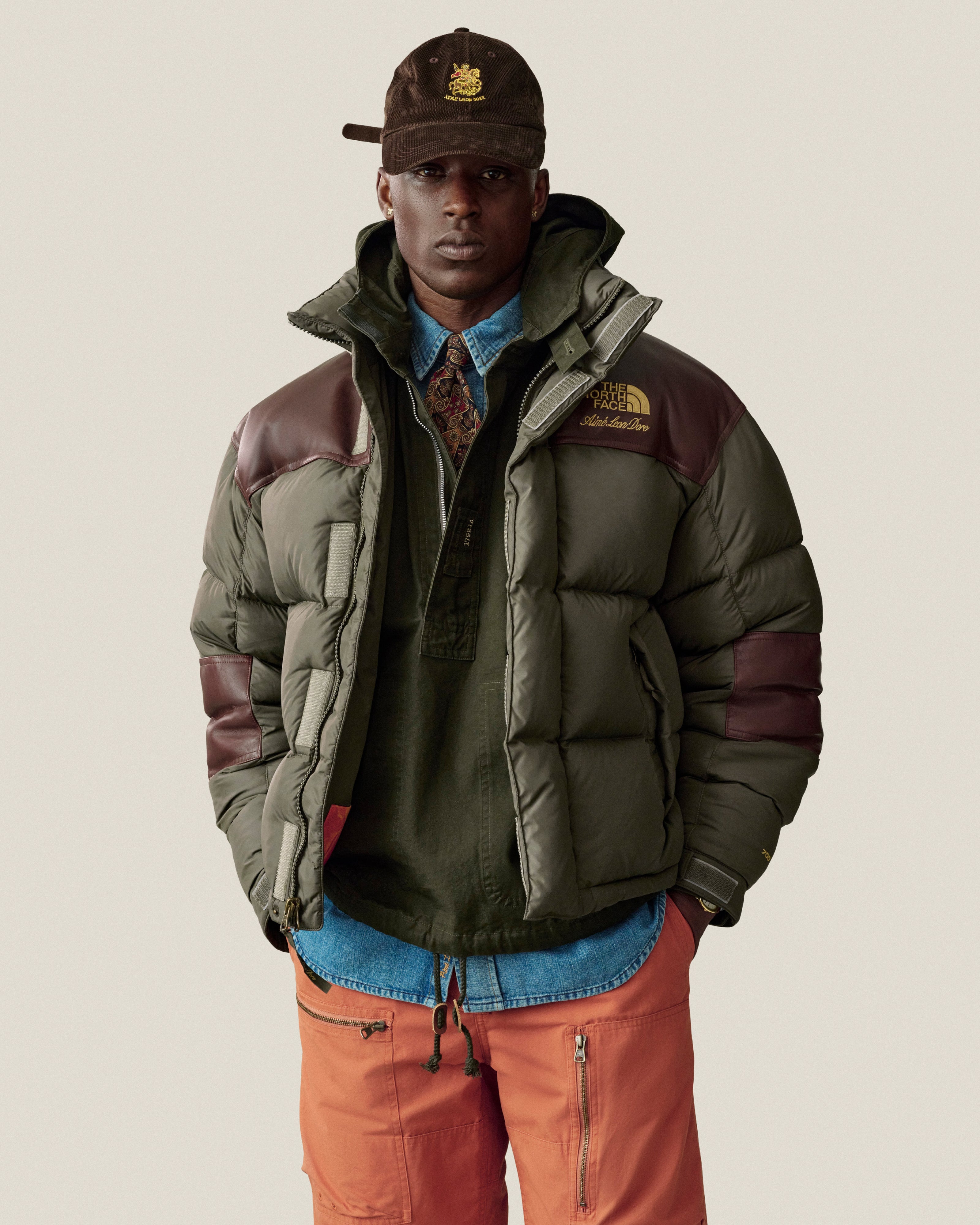 A man in a puffer jacket, Saint George Corduroy Hat, patterned tie, and orange pants stands against a plain background.