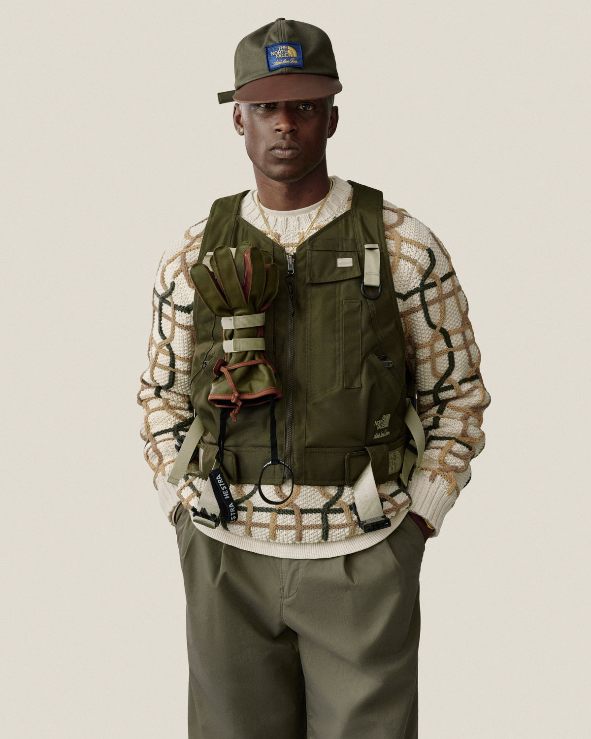 Man in Double Pleated Officer Pant, olive vest, cap, and patterned sweater stands against a plain background.