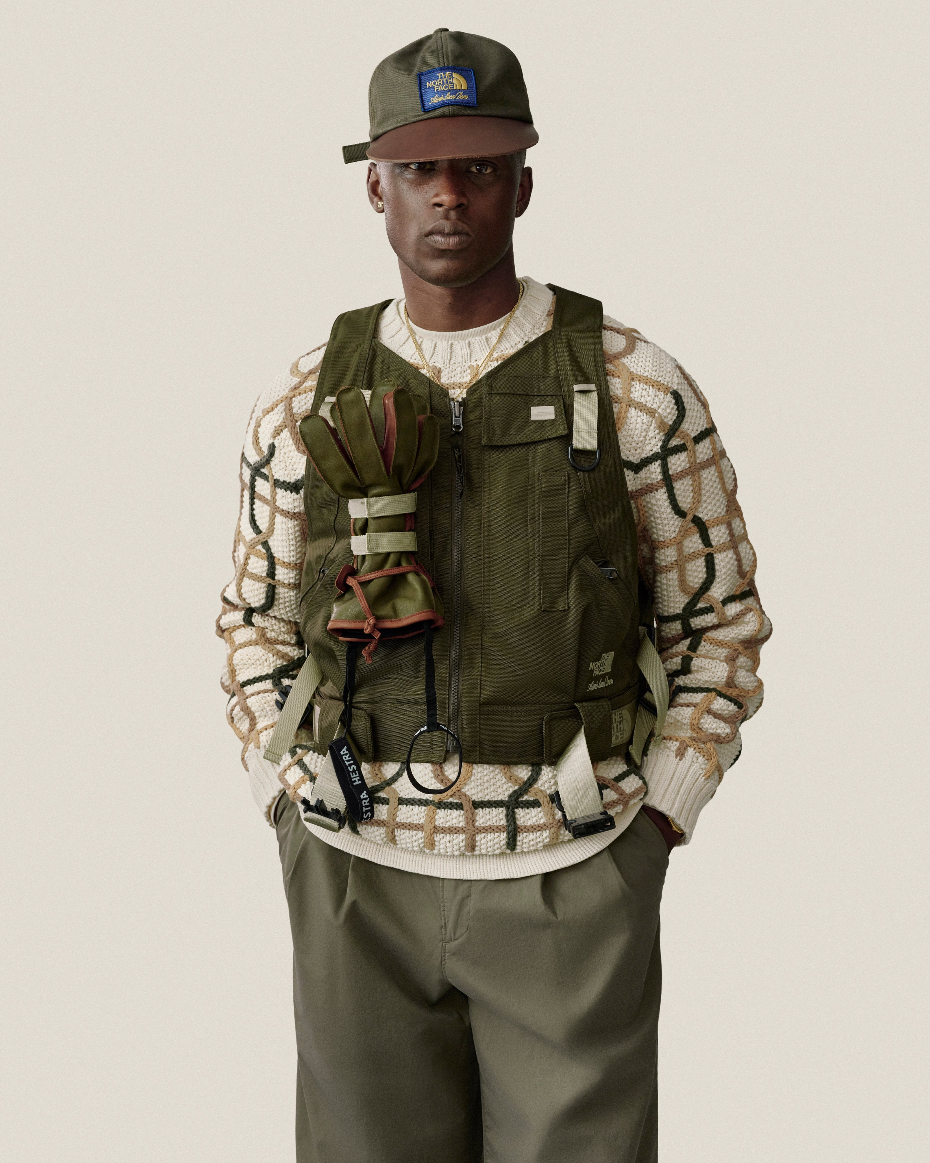 Man in Double Pleated Officer Pant, olive vest, cap, and patterned sweater stands against a plain background.