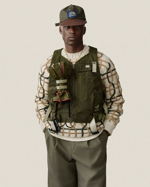 Man in Double Pleated Officer Pant, olive vest, cap, and patterned sweater stands against a plain background.