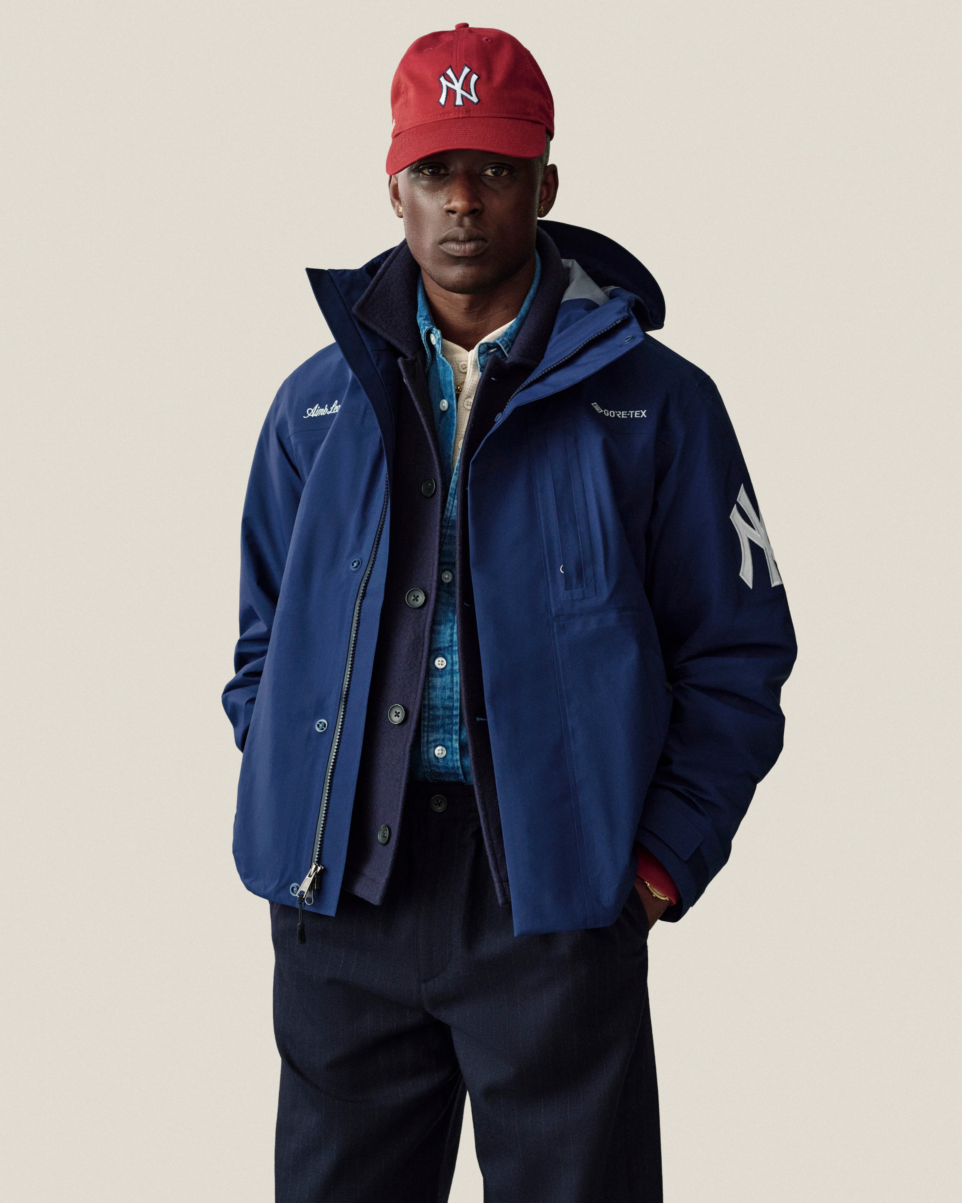 Man in ALD / New Era Yankees Ballpark Hat, NY logo blue jacket, blue shirt, dark pants, on neutral background.