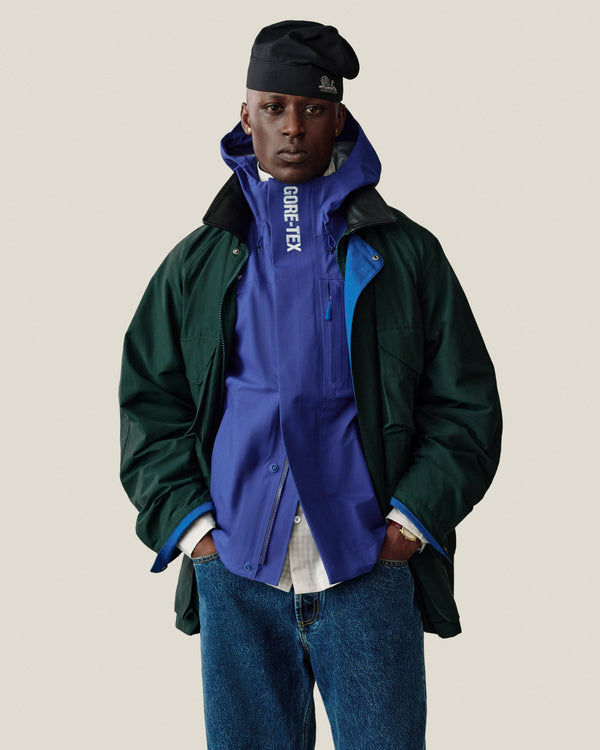 A person in jeans and a black hat stands with hands in pockets wearing a green Dodgers GORE-TEX Rain Delay Jacket.
