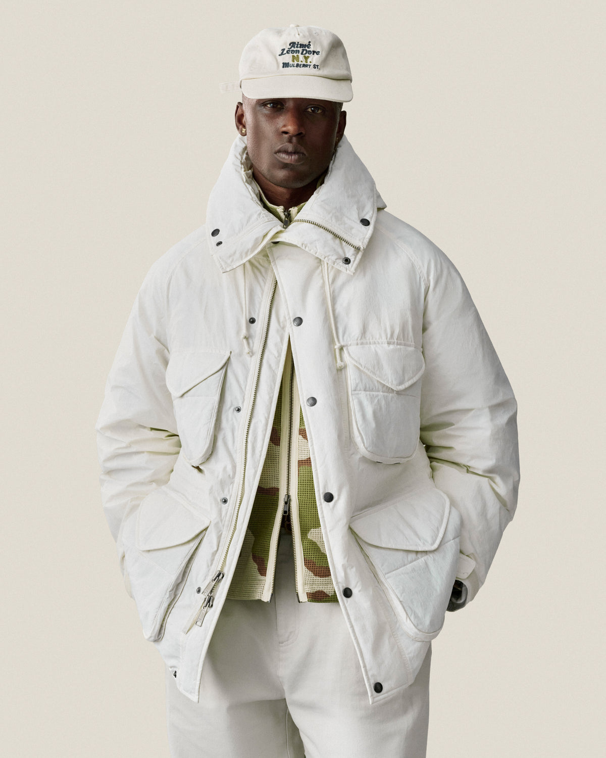 Man wearing a Mulberry Chainstitch Hat, white jacket, and pants, posing with hands in pockets against a light background.