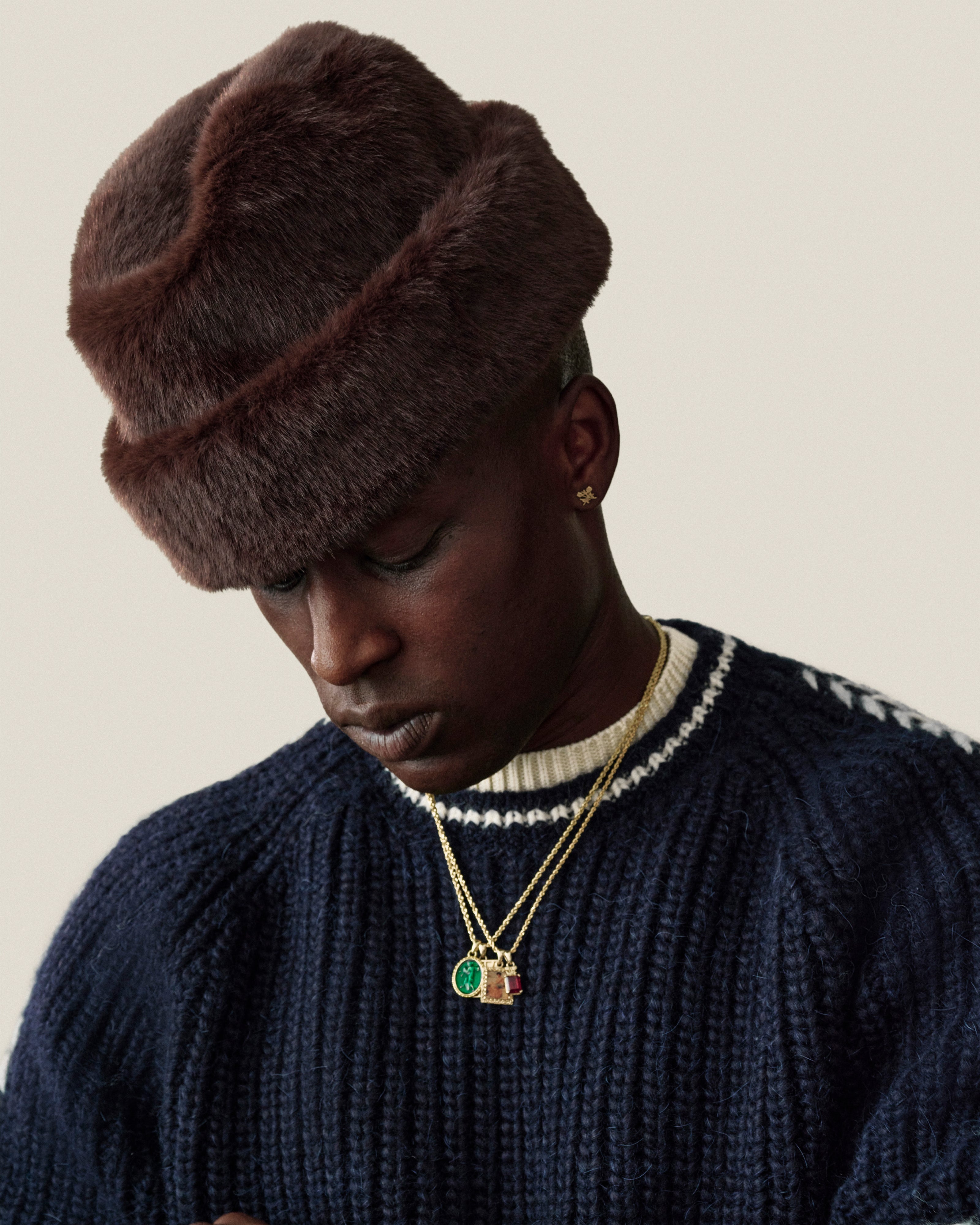 A man wearing a dark brown Fur Envoy Hat and a navy blue ribbed sweater with gold pendant necklaces.