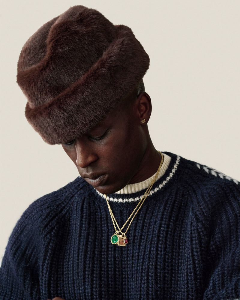 A man wearing a dark brown Fur Envoy Hat and a navy blue ribbed sweater with gold pendant necklaces.