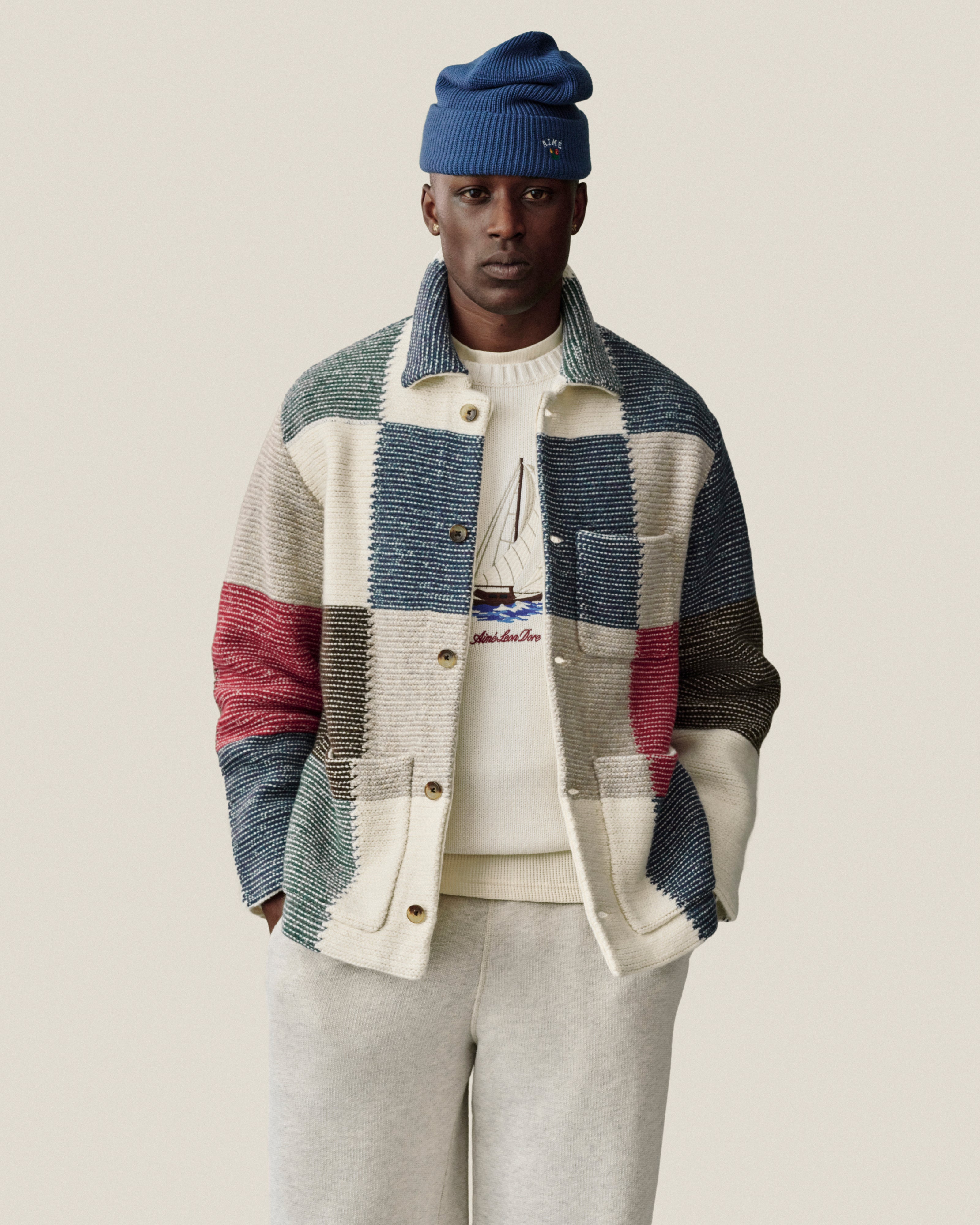 Man in blue beanie, patchwork jacket, sailboat sweater, and Stone Washed Marina Sweatpants stands against plain background.