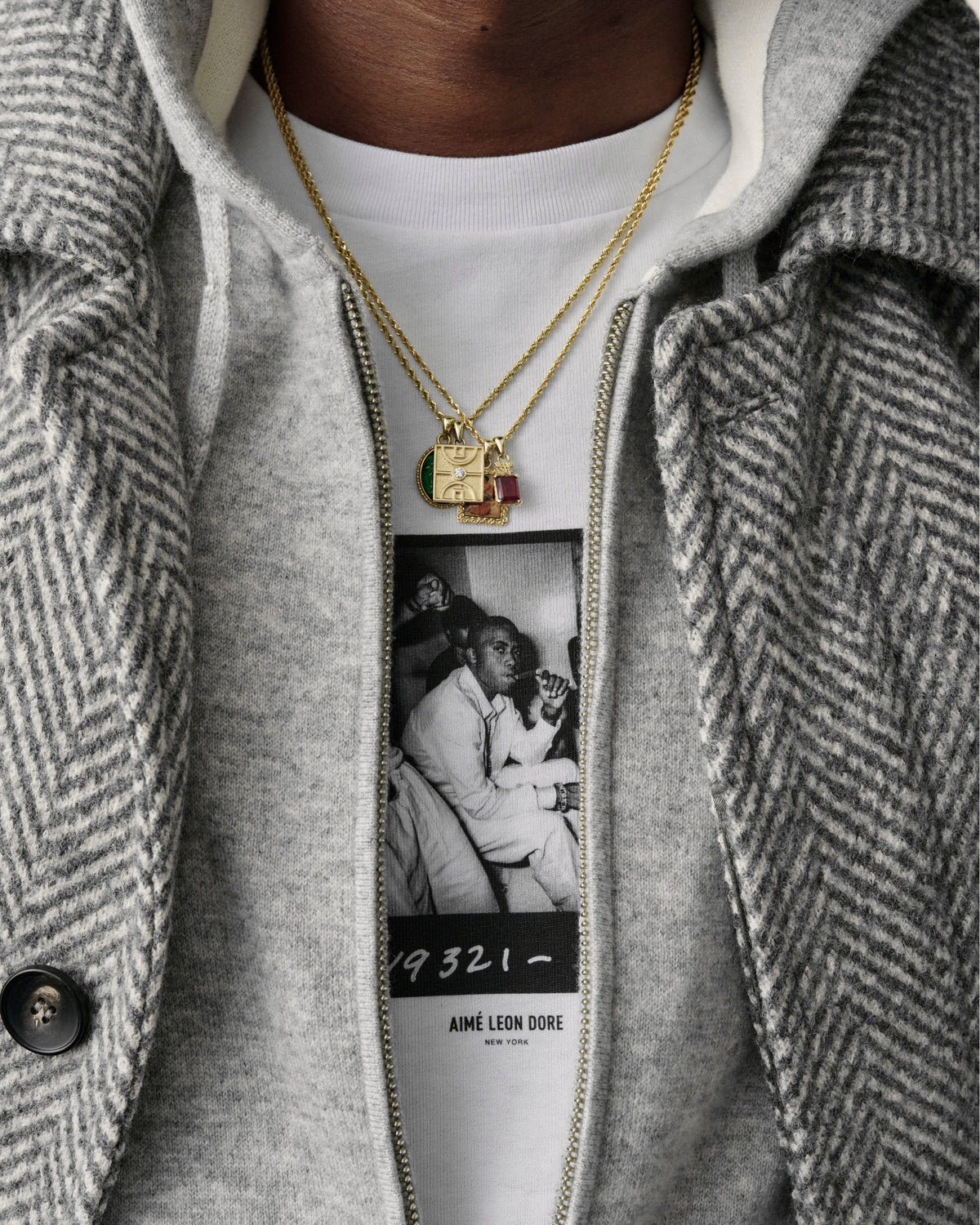 Close-up of a person wearing the 14Kt Gold Diamond Court Pendant, a grey herringbone coat, and a photo-print tee.