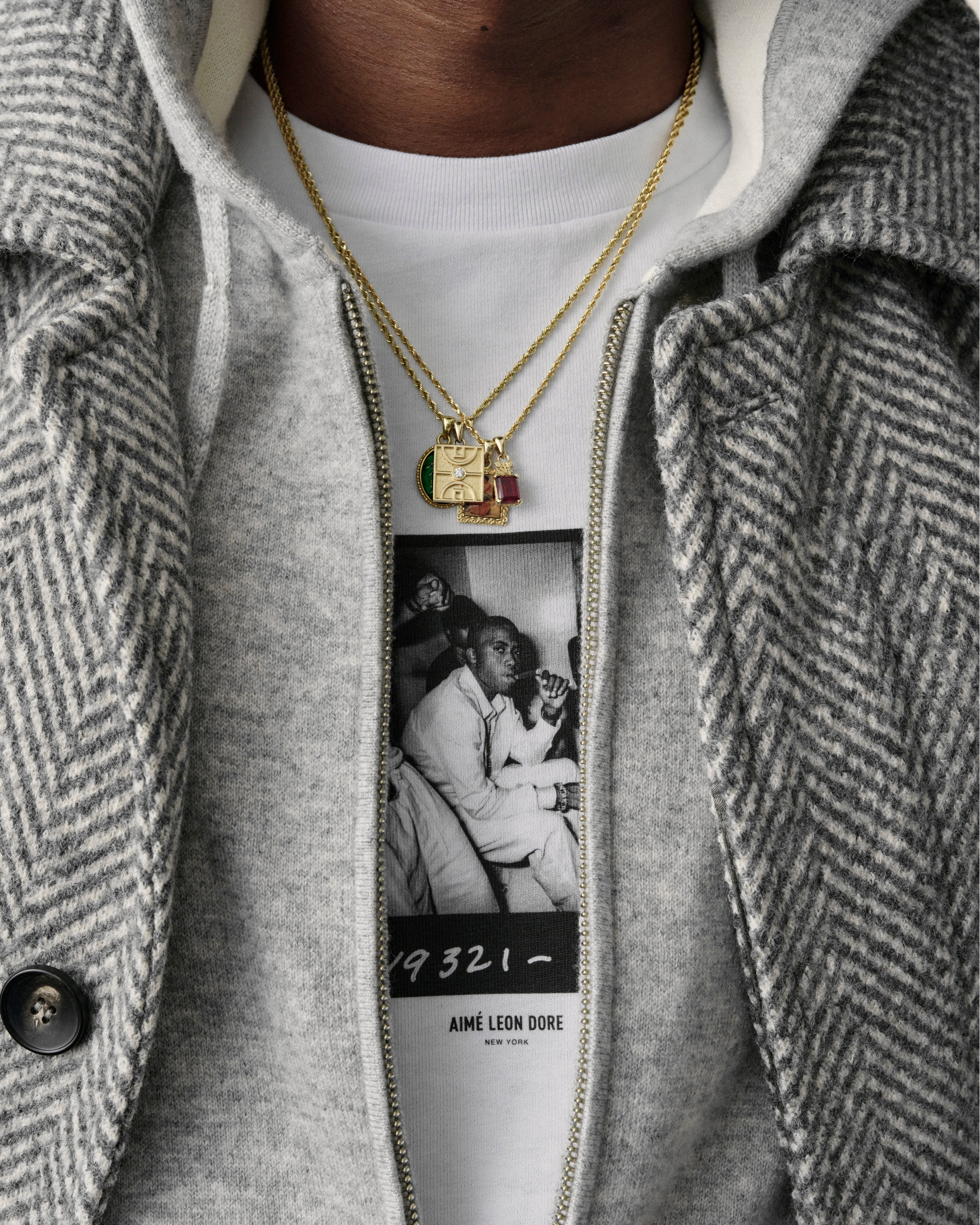 Close-up of a person wearing the 14Kt Gold Diamond Court Pendant, a grey herringbone coat, and a photo-print tee.