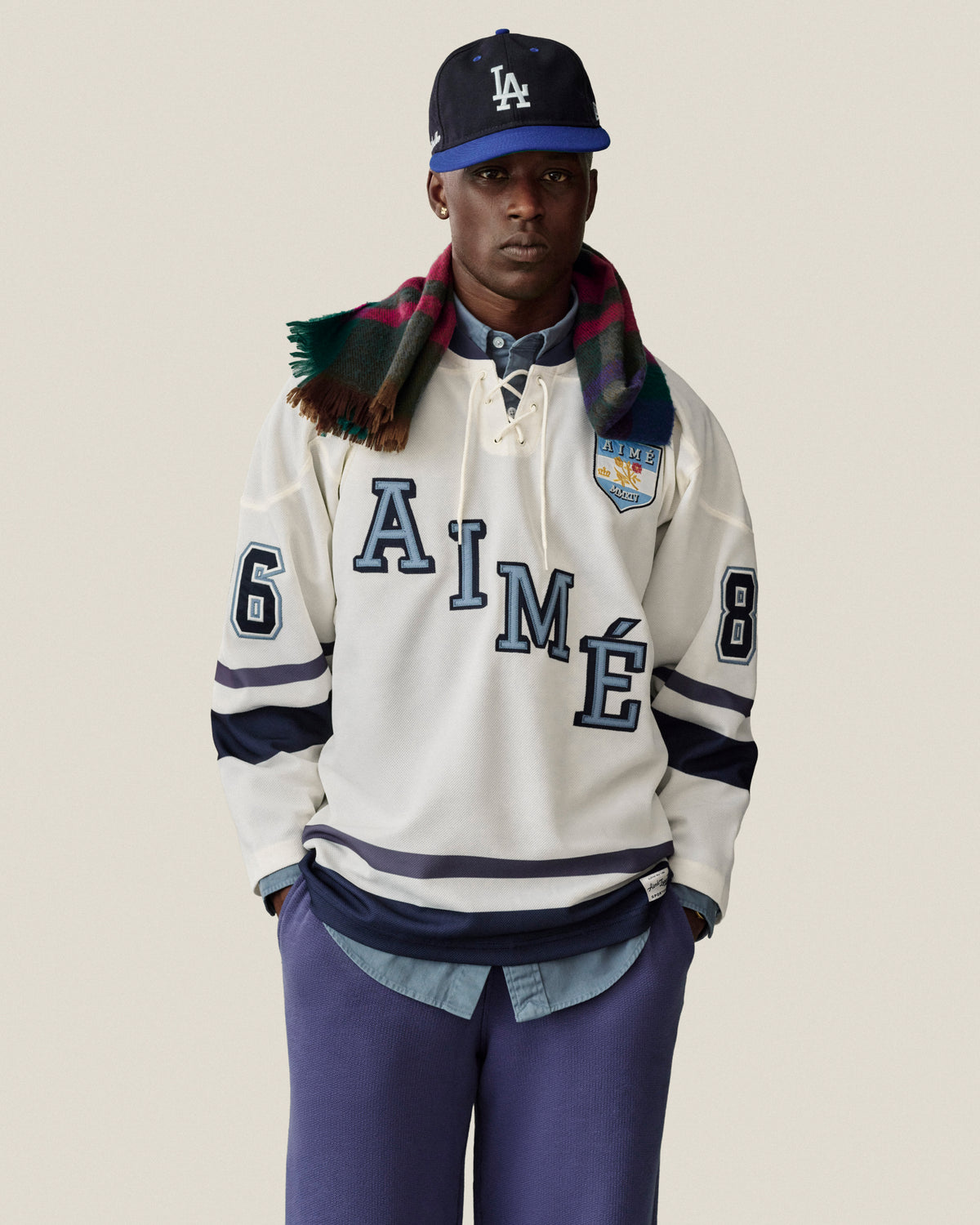 Man in a white hockey jersey, Stone Washed Marina Sweatpants, LA cap, and plaid scarf poses by a neutral backdrop.