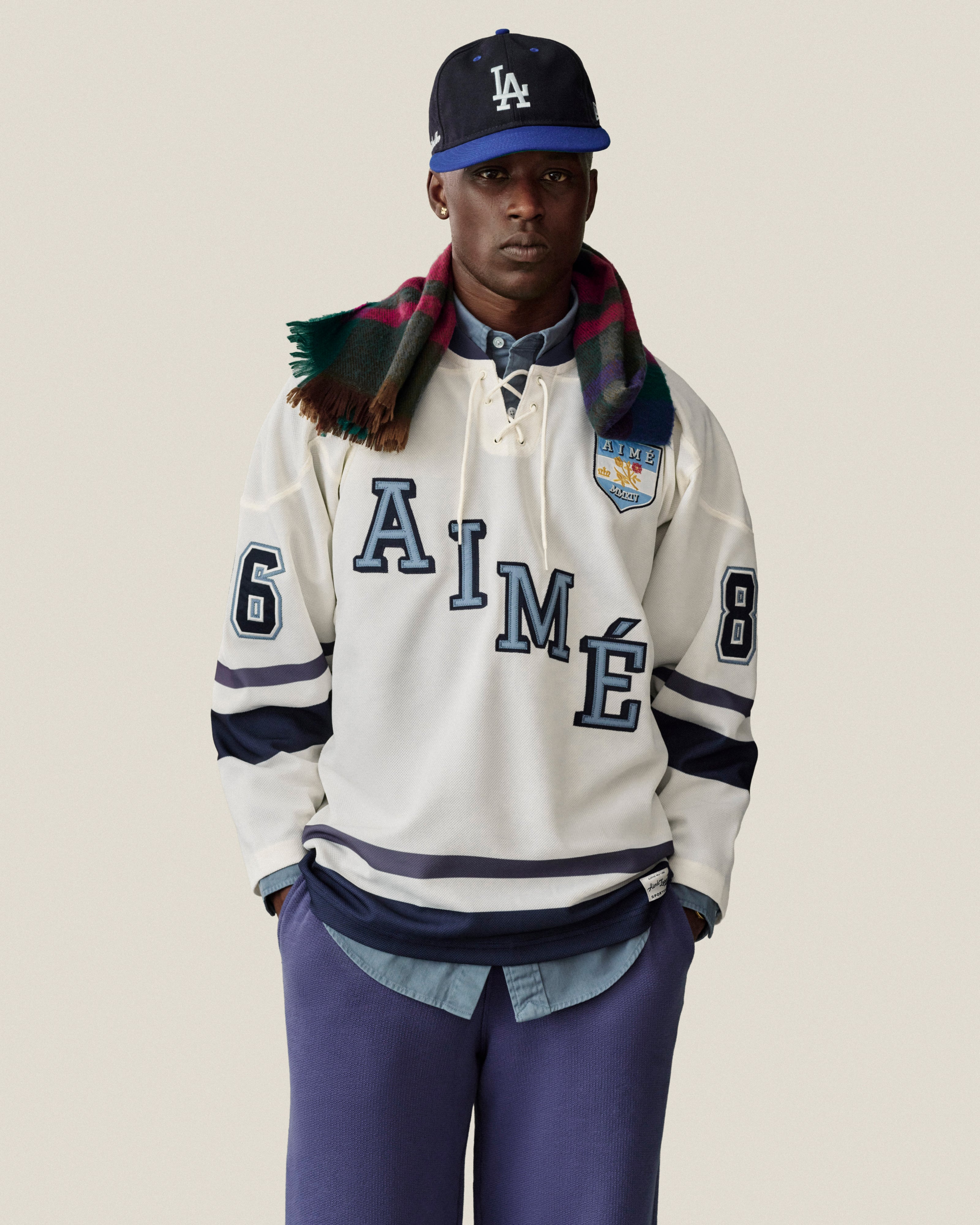 Man in a white hockey jersey, Stone Washed Marina Sweatpants, LA cap, and plaid scarf poses by a neutral backdrop.