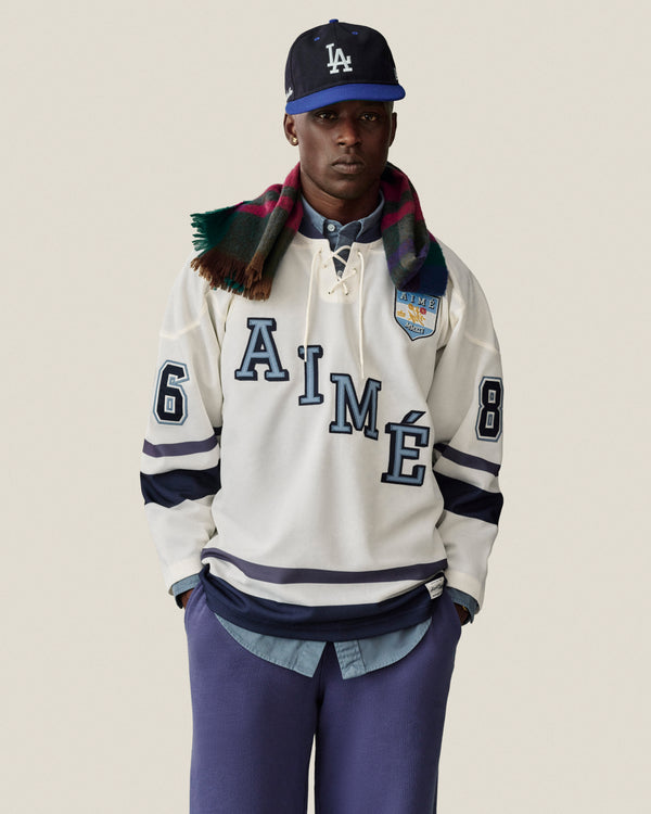 Man in a white hockey jersey, Stone Washed Marina Sweatpants, LA cap, and plaid scarf poses by a neutral backdrop.