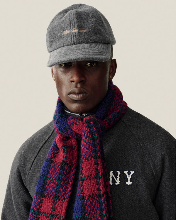 A person wearing a grey Script Fleece Hat with gold script embroidery and a red and blue plaid scarf.
