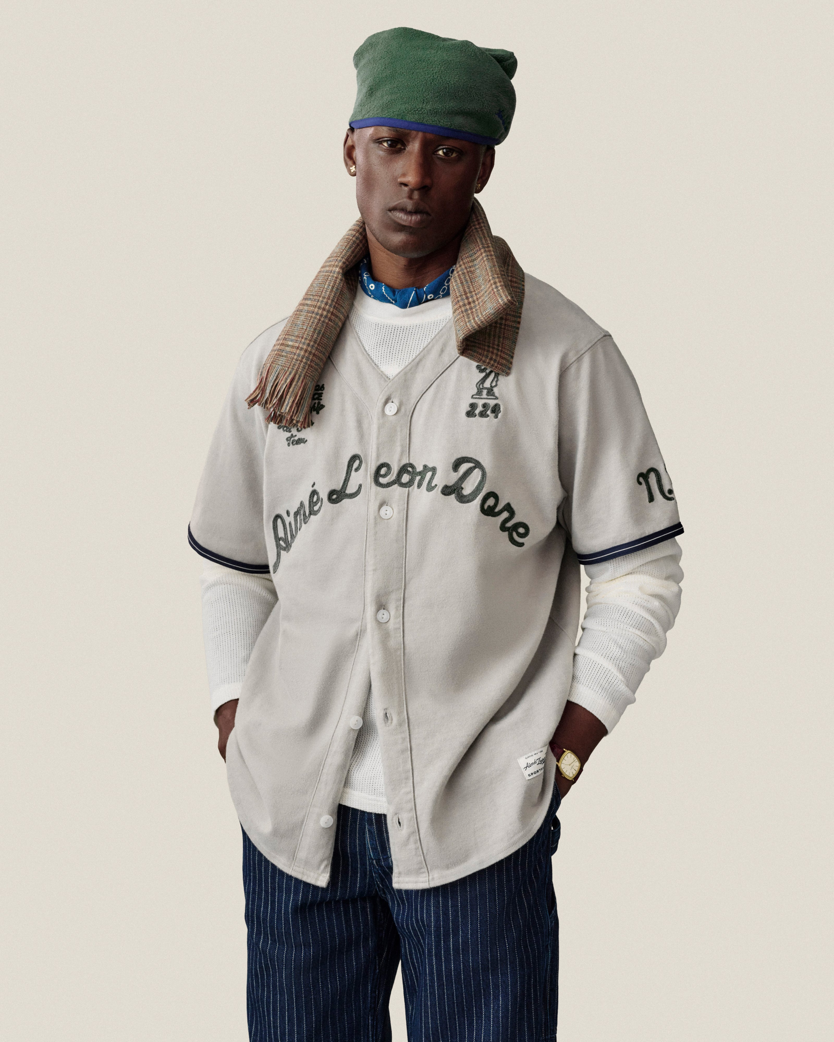 A man in a Long-Sleeve Mesh Football Tee, green beret, scarf, and pinstriped pants stands against a plain background.