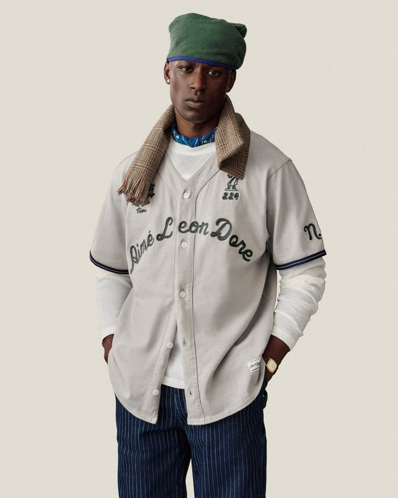 Man in Chalk Stripe Painter Pant, beige jersey, green hat, plaid scarf, and white shirt on plain background.