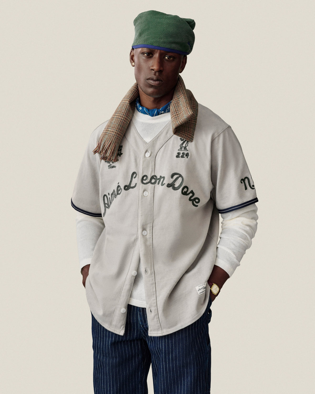 Man in a Mulberry Chainstitch Baseball Jersey, green cap, plaid scarf, and striped pants poses against a plain background.