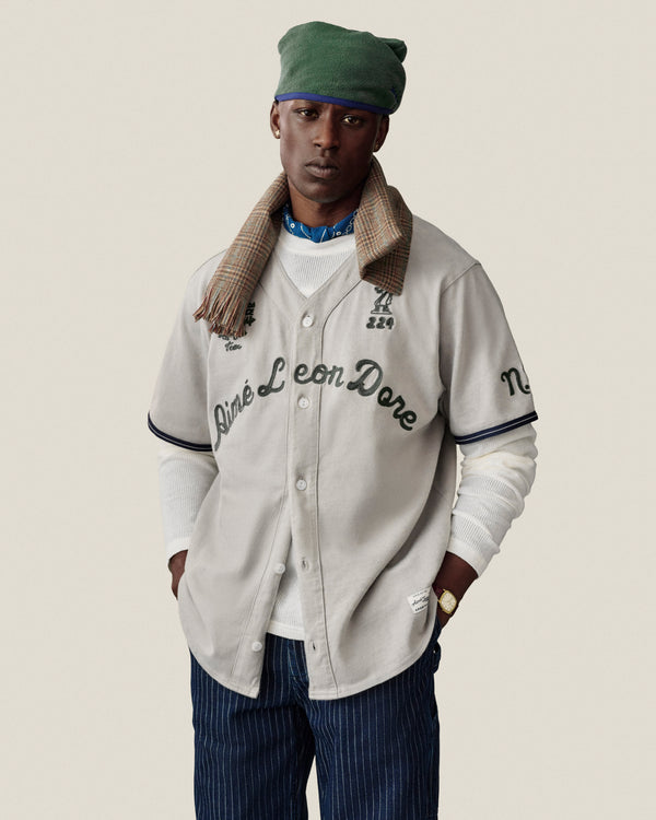 Man in a Mulberry Chainstitch Baseball Jersey, green cap, plaid scarf, and striped pants poses against a plain background.
