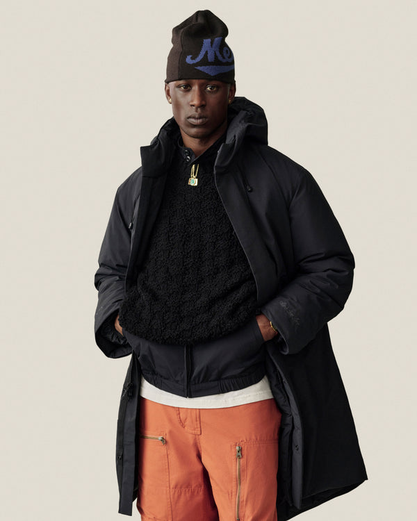 A person wearing a black long coat over a black Fleece Performance Hoodie, orange pants, and a dark beanie.