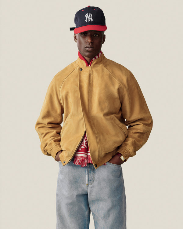 Man in Suede Cafe Racer Jacket, red scarf, blue jeans, and navy NY cap stands before a plain light background.