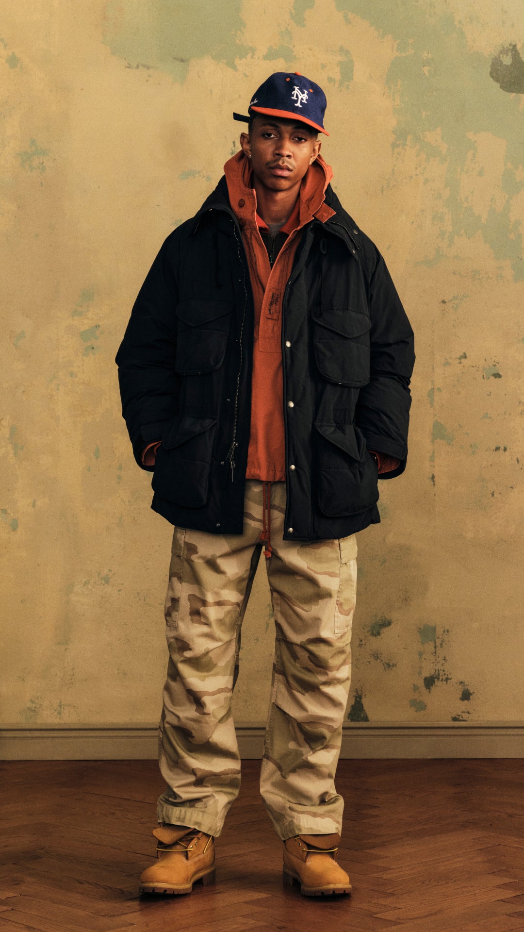 A man stands against a textured beige wall wearing a black puffer jacket over an orange hoodie, beige cargo pants, and tan boots, with a navy baseball cap featuring a white New York Yankees logo.