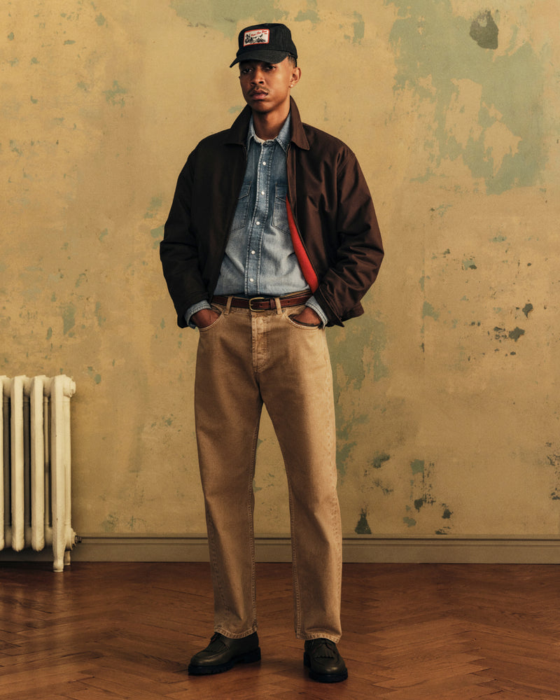 A man wearing an Aimé Retriever Hat, dark jacket, denim shirt, and tan pants.